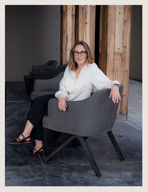 Stacie Jeanjaquet, a creative director, sits comfortably in a modern gray chair, wearing a silk blouse and glasses. The setting features warm wooden beams and a sleek, industrial design.